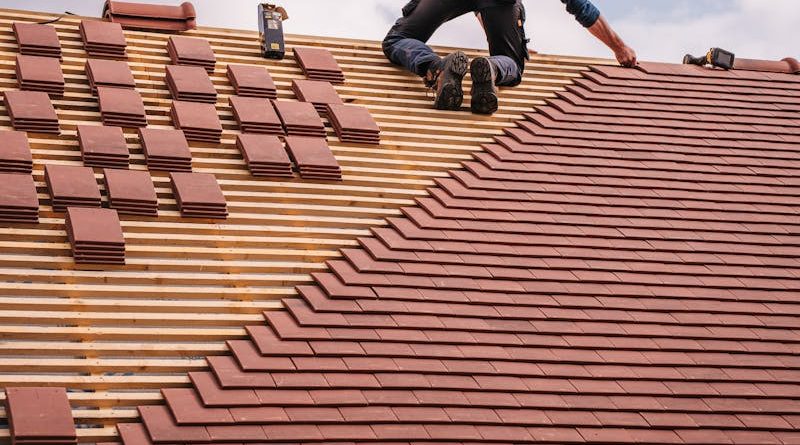 A roofer carefully installs shingles on a new residential roof under a cloudy sky.